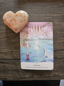 Imrama oracle card beside a peach-colored heart stone on a wooden surface, symbolizing inner guidance and soul exploration