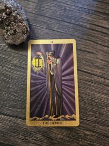 The Hermit tarot card with a glowing lantern and dark robe on a wooden surface beside a sparkling crystal cluster