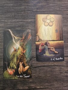 Two tarot cards, The Tower and Five of Pentacles, placed on a wooden surface symbolizing spiritual upheaval and growth through challenge