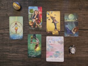 Seven tarot and oracle cards arranged on a wooden surface with crystals and a “Be Fearless” charm, featuring The World, Four of Cups, Page of Swords, The Moon, The Hanged Man, and Trust Your Path