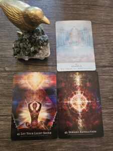 Three oracle cards displayed on a wooden surface with a golden bird statue on a crystal cluster: Atlantis, Let Your Light Shine, and Inward Revolution — representing alignment, inner awakening, and authentic expression.