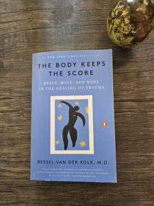 The book The Body Keeps the Score by Bessel van der Kolk, M.D., placed on a wooden surface beside a crystal sphere, representing trauma healing through the connection of brain, mind, and body.