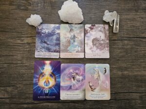 Six oracle cards displayed on a wooden surface surrounded by white crystals, featuring messages including Fortune, Child of the Cosmos, Baby Steps, Divine Masculine, Enlightenment, and Ascension Lift.