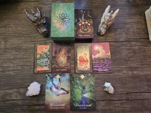 A vibrant tarot and oracle card spread on a wooden surface, featuring cards like Dreamworld, Ace of Embers, The Swan, Four of Trees, Whale Spirit, and Peacock Spirit surrounded by crystals and dragon-shaped stones