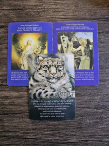 Three oracle cards displayed on a wooden surface. Two Archangel Michael cards show divine light and angelic figures with the messages “Focus Upon Divine and Perfect Health” and “You Created This Situation, and You Have the Power to Change It.” The third card features a snow leopard titled “Self-Reliance,” symbolizing independence, intuition, and personal power.