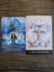 Two oracle cards on a wooden surface — the first features a blue water dragon and a woman holding two chalices labeled “Saga & Sheallah’Shaghthan,” symbolizing wisdom and fluidity, and the second shows a luminous being with white hair and a star crown titled “Trust Your Innocence.”